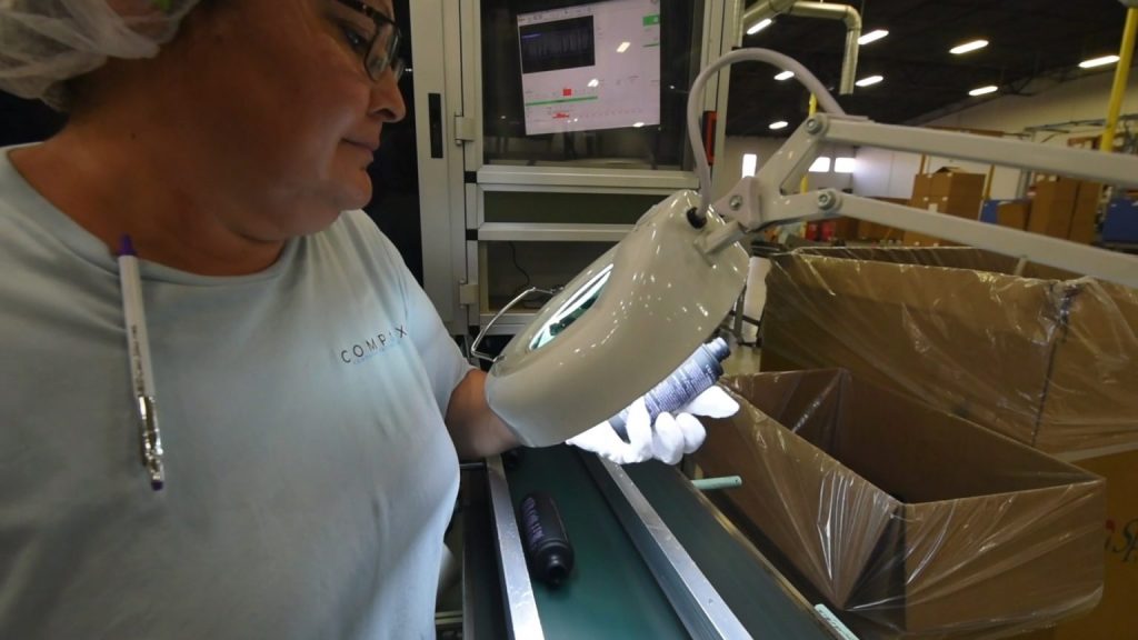A person wearing glasses and a hairnet inspects a small handheld item under a lit magnifying lamp on a conveyor belt in a factory. A computer screen and large cardboard boxes are visible in the background. A person wearing glasses and a hairnet inspects a small handheld item under a lit magnifying lamp on a conveyor belt in a factory. A computer screen and large cardboard boxes are visible in the background.
