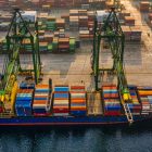 Aerial view of a busy shipping port with a large cargo ship docked and being loaded with colorful stacked shipping containers. The port contains numerous containers organized in rows, and two large cranes are positioned over the vessel, assisting with the loading.