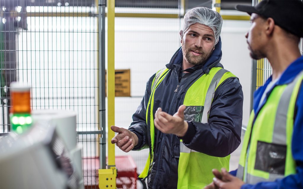Two factory workers in safety vests and hairnets engaging in a discussion on the production floor. One man gestures with his hand while speaking, and the other listens attentively. They stand near industrial equipment enclosed by metal fencing. Two factory workers in safety vests and hairnets engaging in a discussion on the production floor. One man gestures with his hand while speaking, and the other listens attentively. They stand near industrial equipment enclosed by metal fencing.