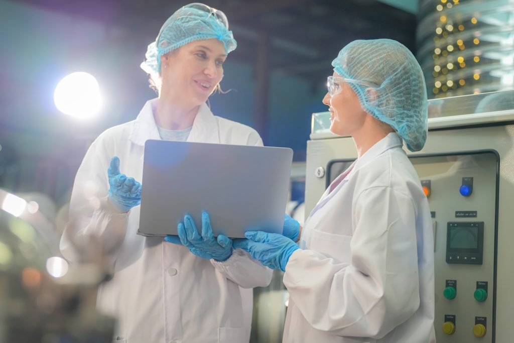 Two scientists wearing lab coats, hairnets, safety glasses, and blue gloves are working in a laboratory. One holds a laptop while they discuss their work. Electronic equipment is visible in the background, suggesting a high-tech research environment. Two scientists wearing lab coats, hairnets, safety glasses, and blue gloves are working in a laboratory. One holds a laptop while they discuss their work. Electronic equipment is visible in the background, suggesting a high-tech research environment.