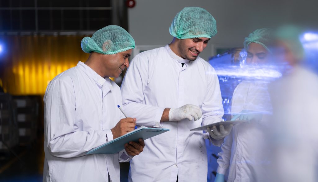 Three people in white lab coats and hairnets stand in a laboratory. Two are holding clipboards and pens, while one points at something on a tablet. They seem to be engaged in a discussion or sharing information. The background is slightly blurred with lab equipment visible. Three people in white lab coats and hairnets stand in a laboratory. Two are holding clipboards and pens, while one points at something on a tablet. They seem to be engaged in a discussion or sharing information. The background is slightly blurred with lab equipment visible.