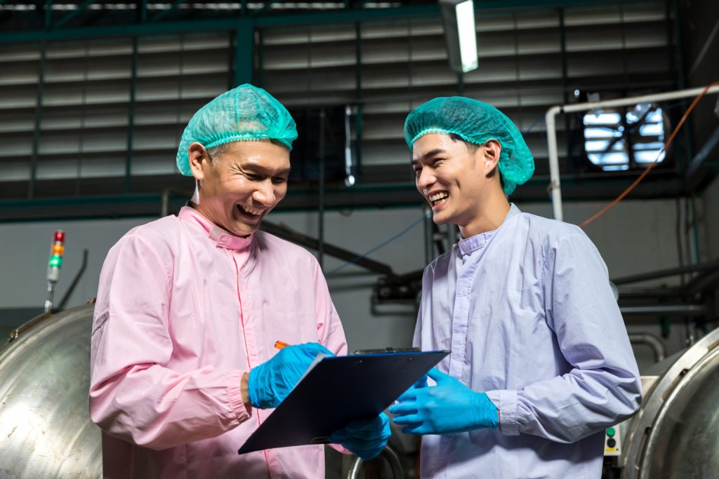 Two smiling industrial workers wearing pink and blue protective clothes, including hairnets and gloves, stand in a factory setting. One is writing on a clipboard, while the other looks on. They appear to be engaged in a friendly conversation. Two smiling industrial workers wearing pink and blue protective clothes, including hairnets and gloves, stand in a factory setting. One is writing on a clipboard, while the other looks on. They appear to be engaged in a friendly conversation.