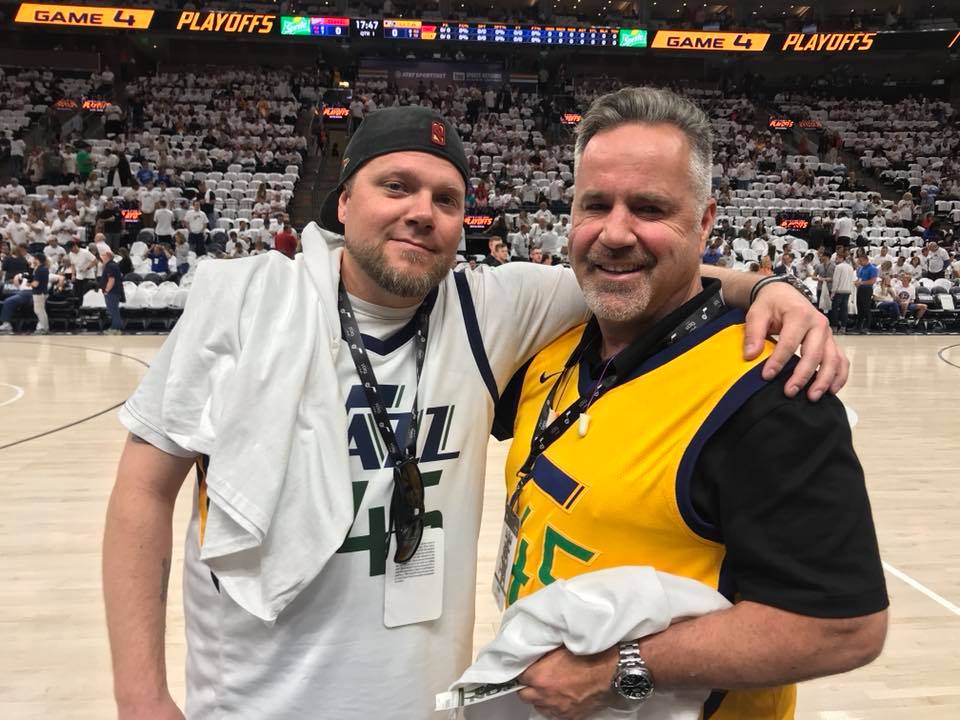 Two smiling men pose on an indoor basketball court during a playoff game. Both are wearing jerseys; the man on the left is in a white Utah Jazz jersey, and the man on the right is in a yellow Utah Jazz jersey. Fans fill the stands in the background. Two smiling men pose on an indoor basketball court during a playoff game. Both are wearing jerseys; the man on the left is in a white Utah Jazz jersey, and the man on the right is in a yellow Utah Jazz jersey. Fans fill the stands in the background.
