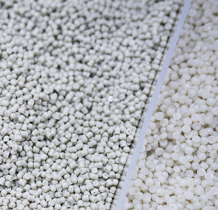 Close-up image of small, white plastic pellets. The pellets are shown partially in a clear container, with some spilling out onto the surface below. The background is blurred, emphasizing the pellets in the foreground.