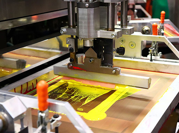 A machine is performing a screen printing process, applying yellow and red ink onto a substrate. The print head moves across the screen, transferring the design. Various mechanical components and clamps are visible, securing the screen in place.