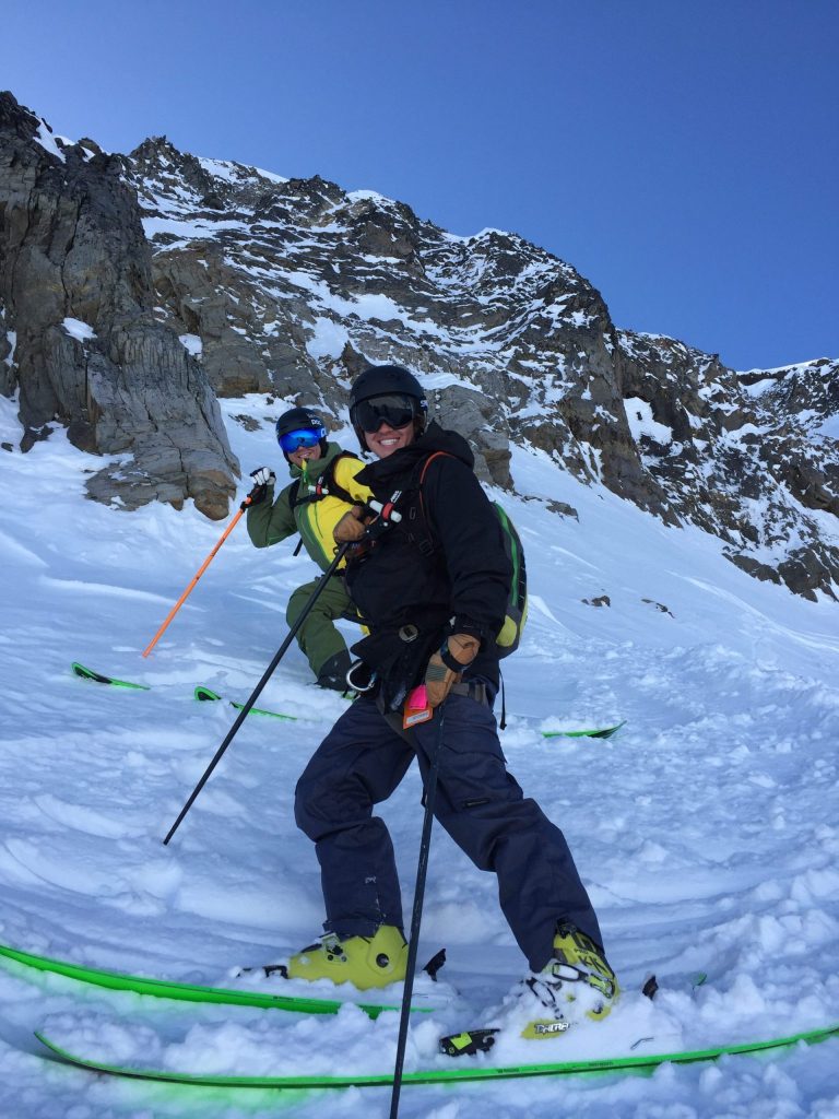 Two skiers dressed in winter gear, including helmets and goggles, pause on a snowy mountain slope for a photo. Both hold ski poles and smile at the camera. The backdrop features rugged, snow-covered rocks beneath a clear blue sky. Two skiers dressed in winter gear, including helmets and goggles, pause on a snowy mountain slope for a photo. Both hold ski poles and smile at the camera. The backdrop features rugged, snow-covered rocks beneath a clear blue sky.