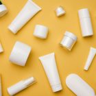 Various white cosmetic containers, including jars, bottles, tubes, and pumps, are scattered randomly on a mustard yellow background. The containers are blank and made of plastic.