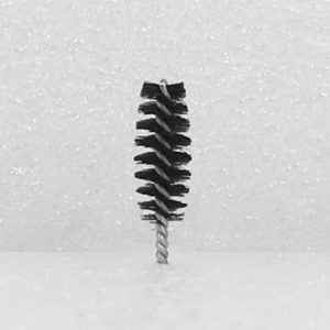 The Nylon Brush Applicator, featuring a small cylindrical design with tightly packed black bristles and a twisted metal handle, stands upright against a white, speckled background. The bristles are arranged in an intricate spiral pattern.