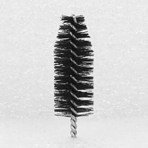 A close-up of a Nylon Brush Applicator standing upright. The applicator features densely packed, spiraled bristles designed for grooming eyelashes or eyebrows. The background is a speckled, light gray surface.