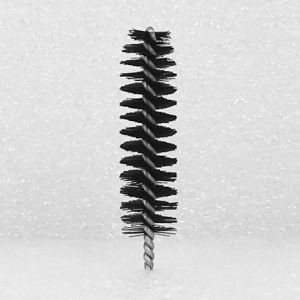 The Nylon Brush Applicator, a small tool featuring black bristles arranged in a spiral pattern, is positioned vertically against a plain, textured background. This brush is commonly used for cleaning gun barrels or small tubes.