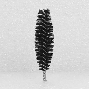 A close-up of the Nylon Brush Applicator, featuring a black, cylindrical brush with dense bristles that resemble a miniature bottle brush. It has a twisted metal handle at the bottom, set against a textured white surface background.