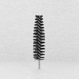 A detailed close-up of the Nylon Brush Applicator, featuring a small cylindrical design with black bristles spiraling around a central metal wire. It stands upright on a textured white surface, with bristles extending outward in all directions.