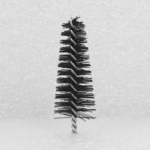 The Nylon Brush Applicator, featuring a narrow and cylindrical black design, stands upright against a white, textured background. The bristles fan out slightly in a spiral pattern around the thin, twisted metal handle at the base.