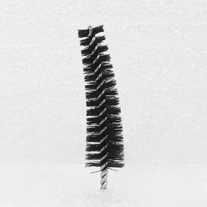A Nylon Brush Applicator, small and black with a twisted wire handle, featuring tightly packed bristles in a cylindrical shape resembling a pipe cleaner, stands upright against a plain, textured background.