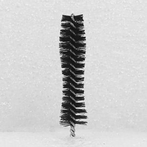 The Nylon Brush Applicator, featuring short black bristles arranged symmetrically and tapering slightly towards the ends, stands upright against a white background. The bristles are attached to a central twisted wire handle in a cylindrical shape.
