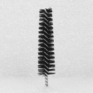 A close-up image of the Nylon Brush Applicator, featuring a cylindrical black brush with tightly packed bristles and a twisted metal stem. The applicator is standing upright against a plain white textured background.