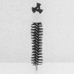 A Nylon Brush Applicator stands vertically against a white background. Above it, a small black bow tie hovers in mid-air. The brush bristles are angled outward, giving it a dense and textured appearance.