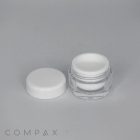 A small, open circular container with a white lid placed beside it, on a plain gray background. The container appears to be made of clear plastic with a white inner lining. The word "COMPAX" is printed in the lower left corner of the image.