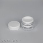 A small, open circular container with a white lid placed beside it, on a plain gray background. The container appears to be made of clear plastic with a white inner lining. The word "COMPAX" is printed in the lower left corner of the image.