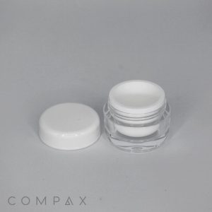 A small, open circular container with a white lid placed beside it, on a plain gray background. The container appears to be made of clear plastic with a white inner lining. The word "COMPAX" is printed in the lower left corner of the image.