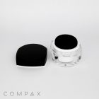 A transparent jar with a black lid containing a cosmetic cream is displayed. The lid is partially removed and placed next to the jar. The background is plain and white, and the word "COMPAX" is faintly visible at the bottom left corner of the image.