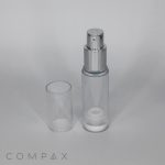 A transparent, cylindrical spray bottle with a silver nozzle and a clear, removable cap placed beside it. The word "COMPAX" is visible at the bottom left corner of the image.