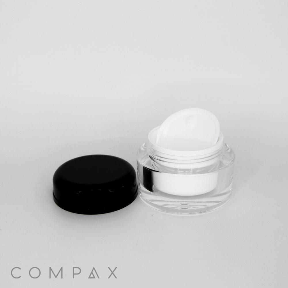 A clear cosmetic jar with a black lid is open, showing a white inner lid and container. The black lid and the white inner lid are placed separately from the jar. The brand name "COMPAX" is visible at the bottom left corner of the image.