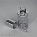 A Round Serum Bottle with a metallic silver cap is open, its translucent dropper placed beside it. The bottle and dropper are set against a plain light background, with the word "COMPAX" visible in the lower left corner of the image.