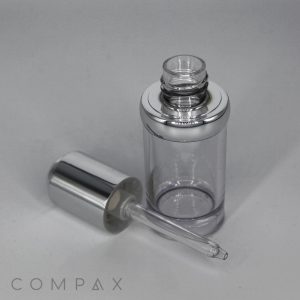 A Round Serum Bottle with a metallic silver cap is open, its translucent dropper placed beside it. The bottle and dropper are set against a plain light background, with the word "COMPAX" visible in the lower left corner of the image.