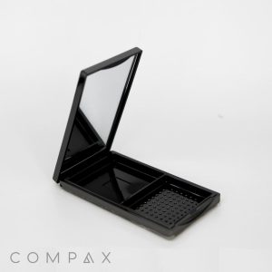 A Single Well Rectangular Compact with Mirror + Brush Well is open, revealing a mirror on the inside of the lid and two empty compartments. The right compartment features a textured surface. The brand name "COMPAX" is displayed at the bottom left. The background is plain white.