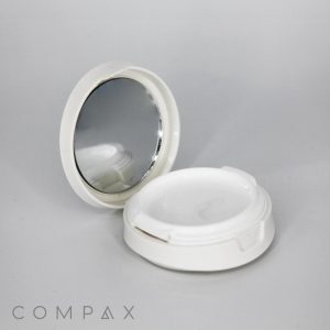 A light gray background sets the stage for a Round Cushion Compact with Mirror, presented in an open position. The round, white compact case features a mirror inside its top lid and an empty compartment in the bottom. The brand name "COMPAX" is clearly visible in the lower left corner.