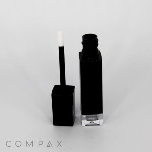 A Square Lip Gloss Bottle featuring a black cap and a clear base is displayed. The container is open with the applicator wand standing beside it, showcasing its white, fuzzy tip. The background is plain white, and "COMPAX" is written in gray at the bottom left corner.