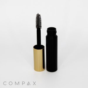 An open Round Mascara Bottle is displayed upright, with the wand partially removed. The mascara wand has a black handle and a spoolie brush. The tube is black with a shiny gold cap. The brand name "COMPAX" is visible in the bottom left corner against a white background.