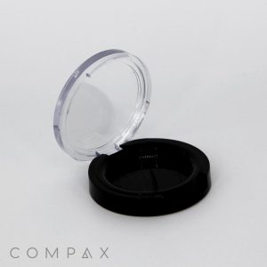 An empty Single Well Round Compact case in black, featuring a clear lid, is resting open against a plain white background. The word "COMPAX" is printed in the bottom left corner.