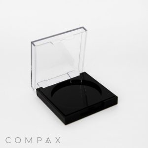 A Square Compact in black with an open clear lid, revealing a circular recess designed to hold makeup. The brand name "COMPAX" is displayed in light gray text at the bottom left corner of the image. The background is plain white.