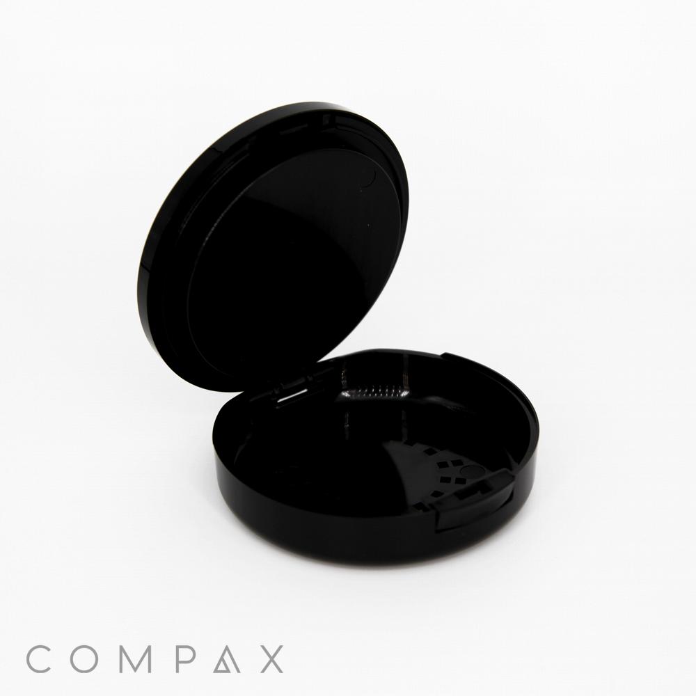 A black, Single Well Round Compact with Mirror features a hinged lid that opens to reveal an empty interior. The compact is set against a white background, with the brand name "COMPAX" displayed in gray text at the bottom left corner of the image.