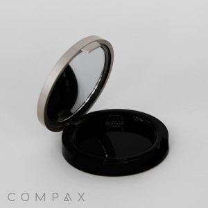 An open Single Well Round Compact in black and silver, featuring a mirror on the inside of the lid. The bottom part of the compact is empty. The word "COMPAX" is printed in gray on the bottom left corner of the image.
