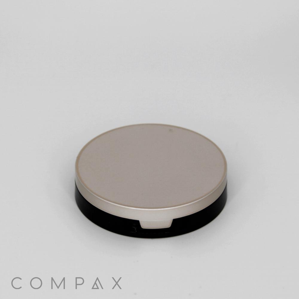 A round, minimalistic beige and black compact case, known as the Single Well Round Compact, is displayed against a plain white background. The word "COMPAX" is visible in light gray text at the bottom-left corner of the image.