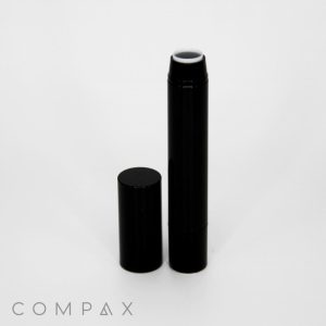 A black cylindrical Round Twist Up Pencil, resembling a large lip balm or glue stick, stands upright with its cap removed and placed next to it. The uncapped pencil reveals a white interior at the top. The word "COMPAX" appears at the bottom left corner.