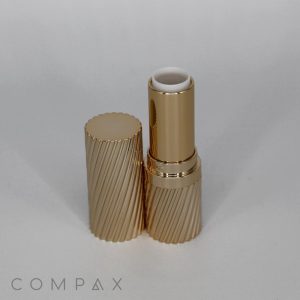 A gold-striped, open tube of Round Textured Lipstick stands upright against a light background. The cap lies next to it on the left. The word "COMPAX" is visible in the bottom left corner of the image. The lipstick itself appears to be unused.
