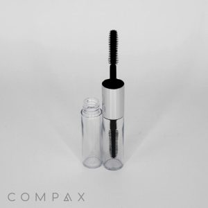 A clear open bottle of Dual Round Mascara with a metallic silver cap and black applicator wand is placed on a light gray background. The cap is removed and standing upright beside the bottle. The word "COMPAX" is visible in gray text at the bottom left corner.
