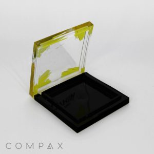 A Single Well Square Compact with a hinged lid, featuring yellow edges and a transparent plastic design. The lid is partially open, revealing an empty black interior. The word "COMPAX" is printed in gray at the bottom left corner of the image.