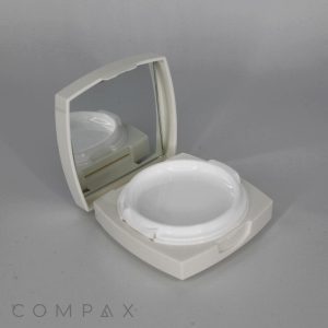 An open Square Cushion Compact features a small mirror inside the lid and an empty circular compartment for makeup or powder. The case is light-colored, with the word "COMPAX" visible in the bottom left corner of the image.