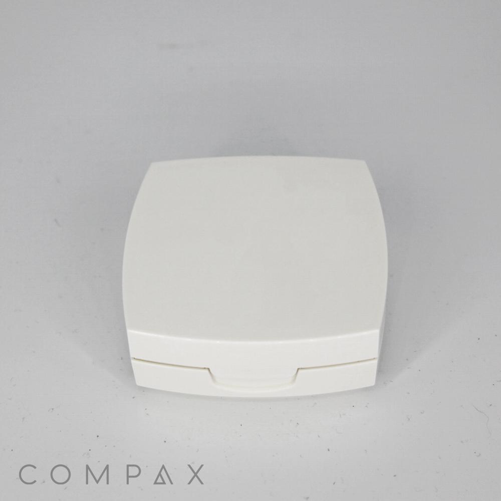 A white, square "Square Cushion Compact" case with a subtly curved lid is centered against a plain background. The brand name "COMPAX" is visible in light gray text at the bottom left corner of the image.