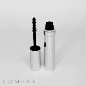 A Round Mascara Bottle in silver stands open with its black brush applicator leaning against it. The minimalistic packaging is clean and modern, showcasing the brand name "COMPAX" at the bottom left corner of the image.