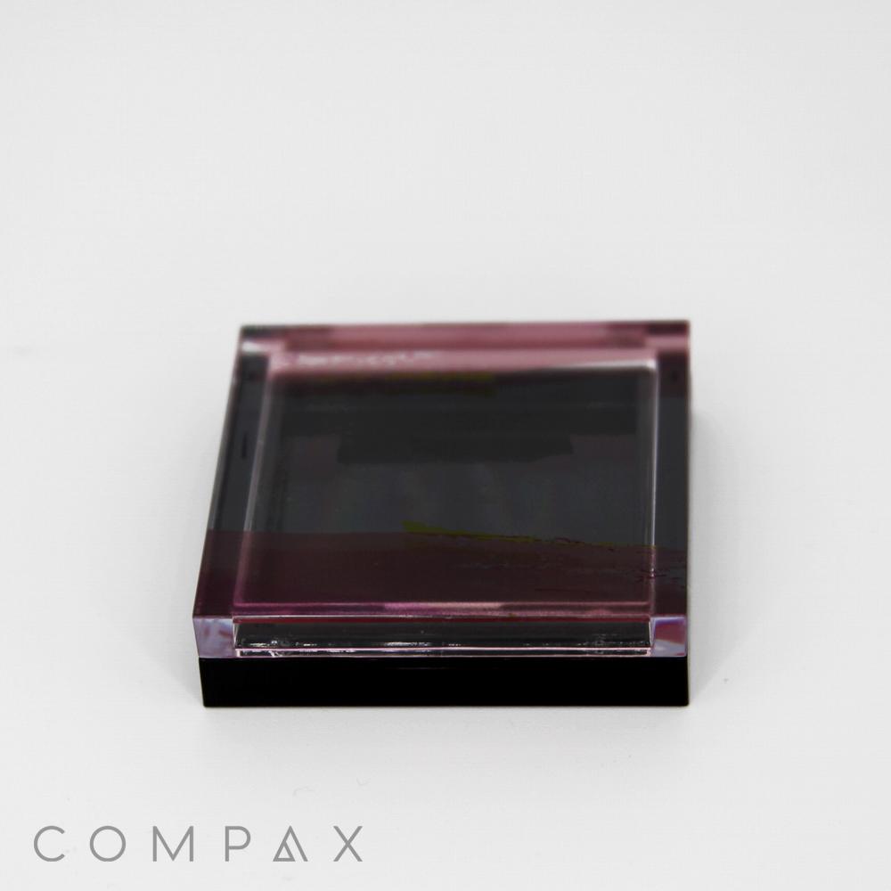 A Single Well Square Compact, transparent with a slight tint, closed shut. It features sleek and minimalist packaging with a subtle black gradient. The brand name 'COMPAX' is written in light gray letters at the bottom left corner of the image on a white background.