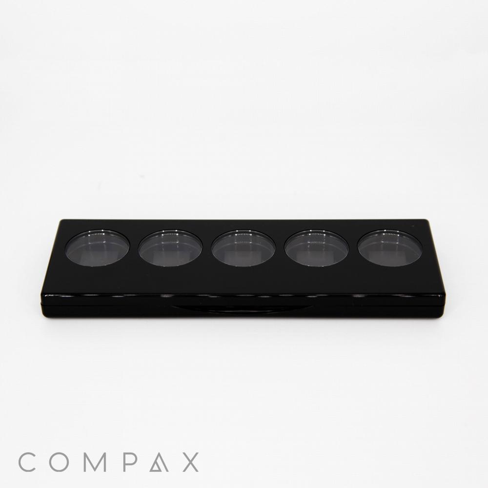 A sleek black 5 Well Rectangular Compact with Windows features five circular compartments arranged in a horizontal row. The brand name "COMPAX" is displayed in small, gray text at the bottom left corner of the image, set against a plain white background.