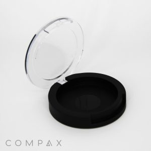 A Single Well Round Compact case, featuring a round, black design and a transparent lid that opens easily, is showcased against a white background. The brand name "COMPAX" is elegantly written in grey letters at the bottom left corner.