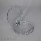 A clear, circular compact case with a hinged lid, identified as the "Single Well Round Compact," is open, revealing its empty interior. The case sits against a plain, light gray background, and the brand name "COMPAX" is visible in the bottom left corner of the image.