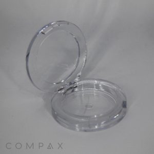 A clear, circular compact case with a hinged lid, identified as the "Single Well Round Compact," is open, revealing its empty interior. The case sits against a plain, light gray background, and the brand name "COMPAX" is visible in the bottom left corner of the image.